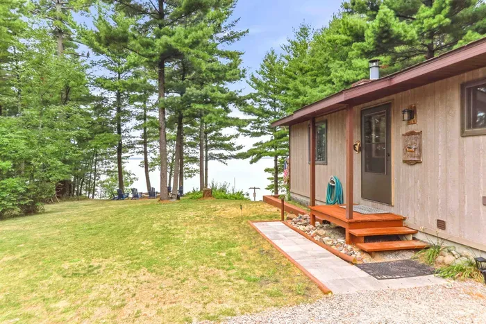 Step up to your welcoming cabin entrance framed by towering pines and sparkling lake views just beyond the lawn. A paved walkway leads directly to the covered entryway, while the expansive grassy yard features an outdoor seating area with Adirondack chairs