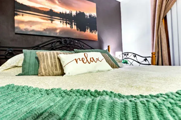 Sink into pure comfort with plush bedding, sage green throw pillows, and a cheerful "Relax" accent pillow. Framed by a scenic sunset canvas above the wrought-iron bed frame, this serene haven is crafted for ultimate rest and rejuvenation.