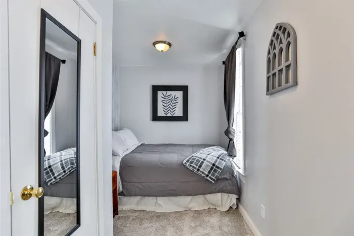 A small, bright bedroom with a centered bed, gray and white bedding, framed wall art, a narrow window with curtains, a wall mirror on the closet door, and simple decorative accents creating a clean, cozy feel.