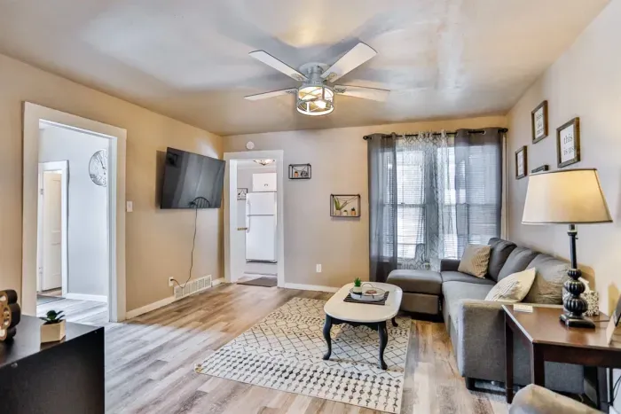 Cozy, sunlit living room with a modern sectional sofa, wall-mounted TV, and warm neutral tones, featuring wood-style floors, a ceiling fan, and an open view into the kitchen.