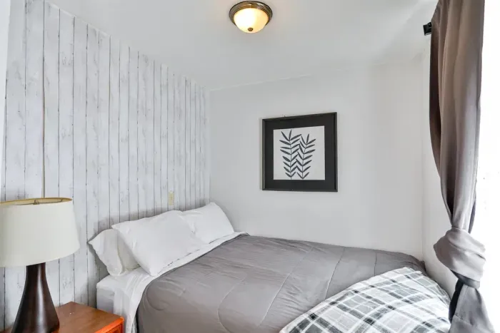 A cozy, minimalist bedroom with a neatly made bed, light gray and white bedding, a wood-panel accent wall, framed leaf artwork, and soft natural light coming through a curtained window.