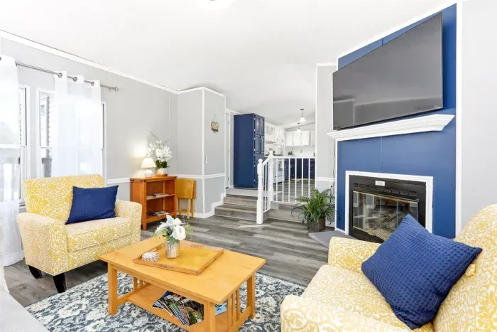 Bright and stylish living space with modern décor, featuring cozy yellow patterned chairs, a blue-accented fireplace with mounted TV, and an open layout leading to a fully equipped kitchen. Perfect for relaxing and entertaining.