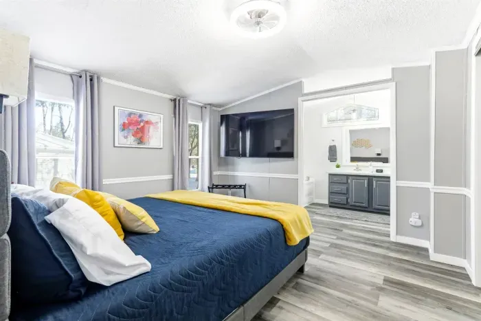 Bright and modern bedroom with a cozy queen-size bed, soft linens, and a calming blue-and-yellow accent palette. Features a wall-mounted smart TV, large windows with plenty of natural light, and sleek wood-style flooring.