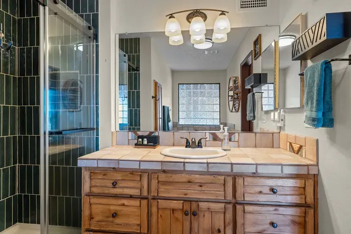 This charming bathroom features a rustic wooden vanity, a tiled countertop, and a walk-in shower with dark green accents. Bright lighting and fresh towels make it comfortable and inviting for guests.