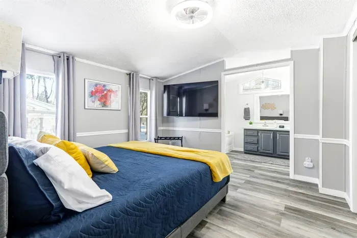 Bright and modern bedroom with a cozy queen-size bed, soft linens, and a calming blue-and-yellow accent palette. Features a wall-mounted smart TV, large windows with plenty of natural light, and sleek wood-style flooring.