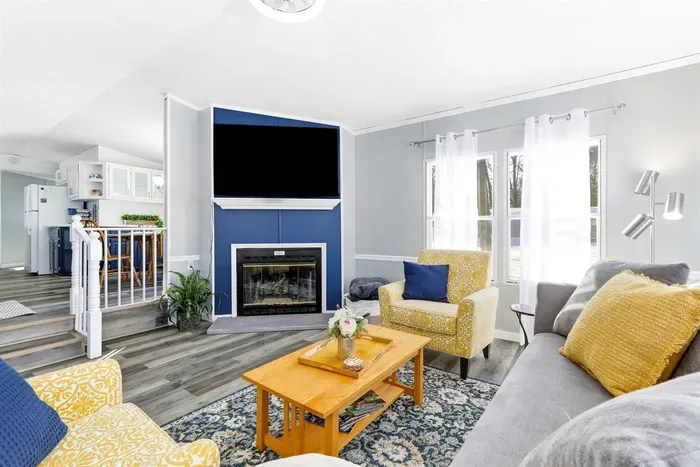 Stylish living space with yellow patterned chairs, a blue-accented fireplace with TV, and an open layout to a modern kitchen, perfect for relaxing or entertaining.