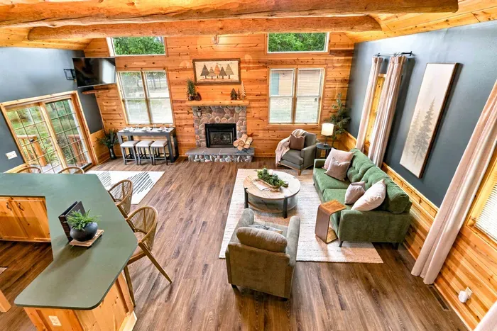 Experience an open-concept layout featuring a stone fireplace, wall-mounted flat-screen TV, spacious kitchen island bar, and soaring wood-beamed ceilings.