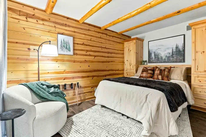 Rest comfortably on a plush bed flanked by custom storage wardrobes, with a soft accent chair set beneath warm, exposed log beams.