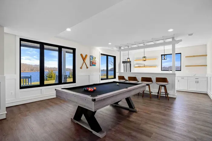 This spacious game room features a comfortable seating area with a modern fireplace, a pool table, ping-pong table, foosball table, shuffleboard, and a well-equipped kitchenette, all with stunning lake views.