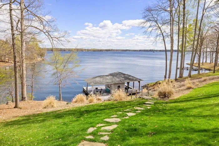 A serene lakeside view with a boat dock and a walking path leading through a grassy lawn flanked by a canvas of trees.