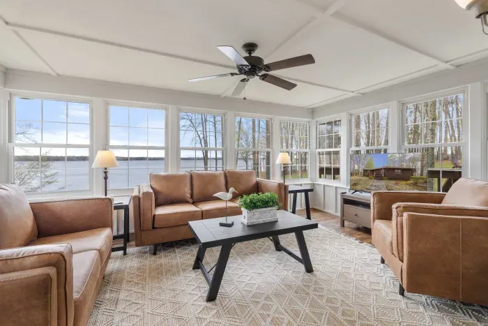 A cozy and bright sunroom furnished with leather sofas and a coffee table, boasting panoramic views of the lake through its many windows.