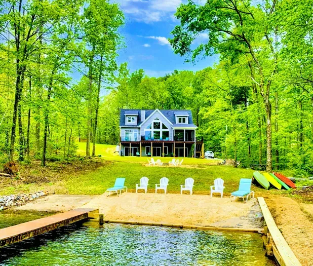 Spacious lakefront home with a private sandy beach, dock, and plenty of outdoor seating surrounded by lush trees