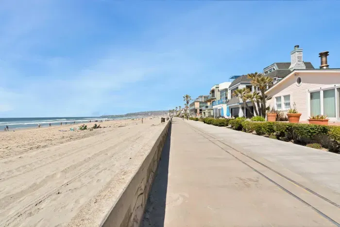 A scenic beachfront promenade lined with houses on one side and a wide, sandy beach on the other stretches into the distance under a clear blue sky.