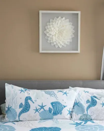 A bedroom features a beige wall with a framed white floral art piece above a gray headboard, and blue and white sea-themed bedding with seahorse, fish, and starfish patterns.